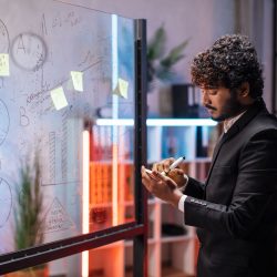 Focused male employee writing on colorful sticky notes on office glass board, developing business project, using tablet pc. Startup plan, people and technologies concept